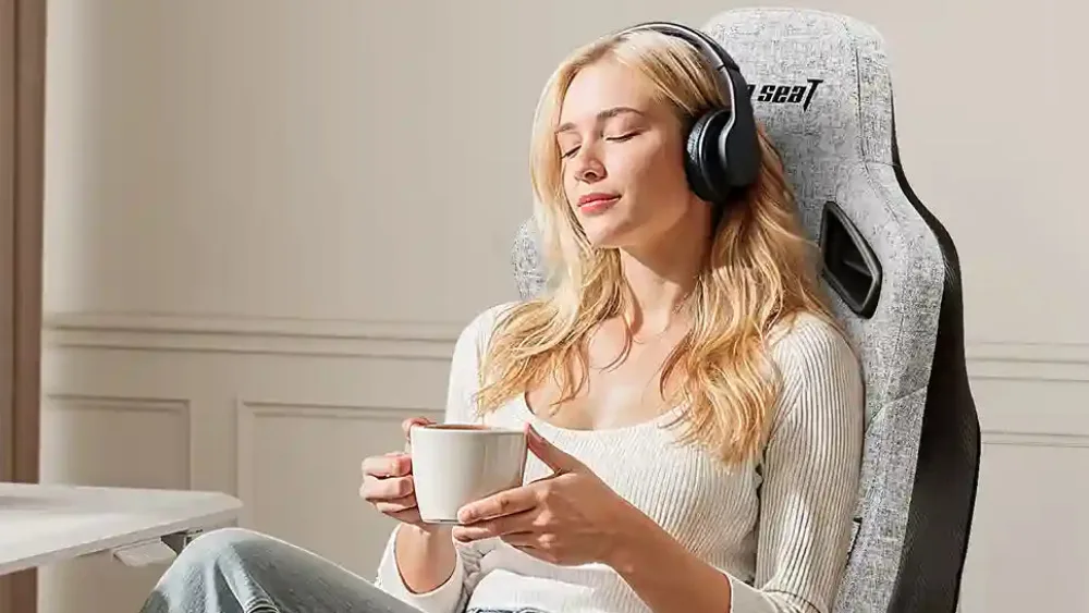 Девушка в наушниках с чашкой отдыхает в кресле AndaSeat Kaiser 3E в серой льняной ткани