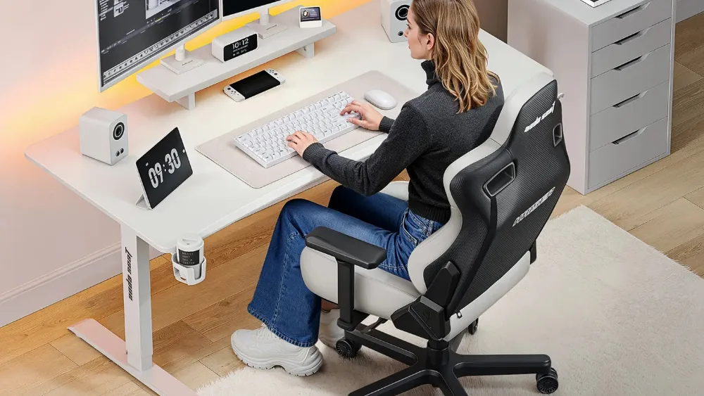 Qiz ikki monitorli oq AndaSeat Xtreme stolida ishlayapti, yonida balandlik displeyli pult