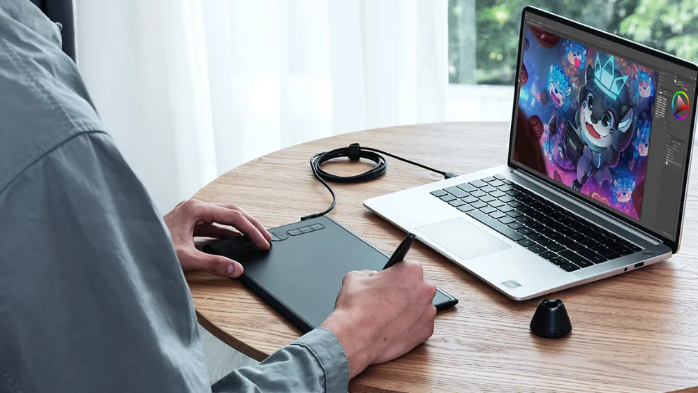 Noutbuk yonidagi Huion Inspiroy H580X: qoʻl planshetda pero bilan chizadi, rasm noutbuk ekranida paydo boʻladi