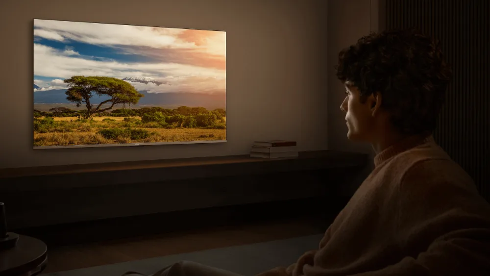 Kechqurun xira yorugʻda tomoshabin Xiaomi TV A 55 ni koʻrmoqda, ekranda quyosh botayotgan savanna